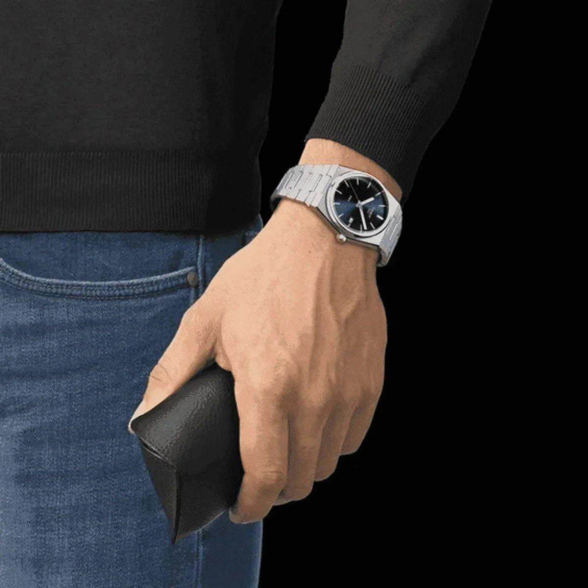 Man wearing silver blue dial watch with metal bracelet, holding black case, jeans and sweater